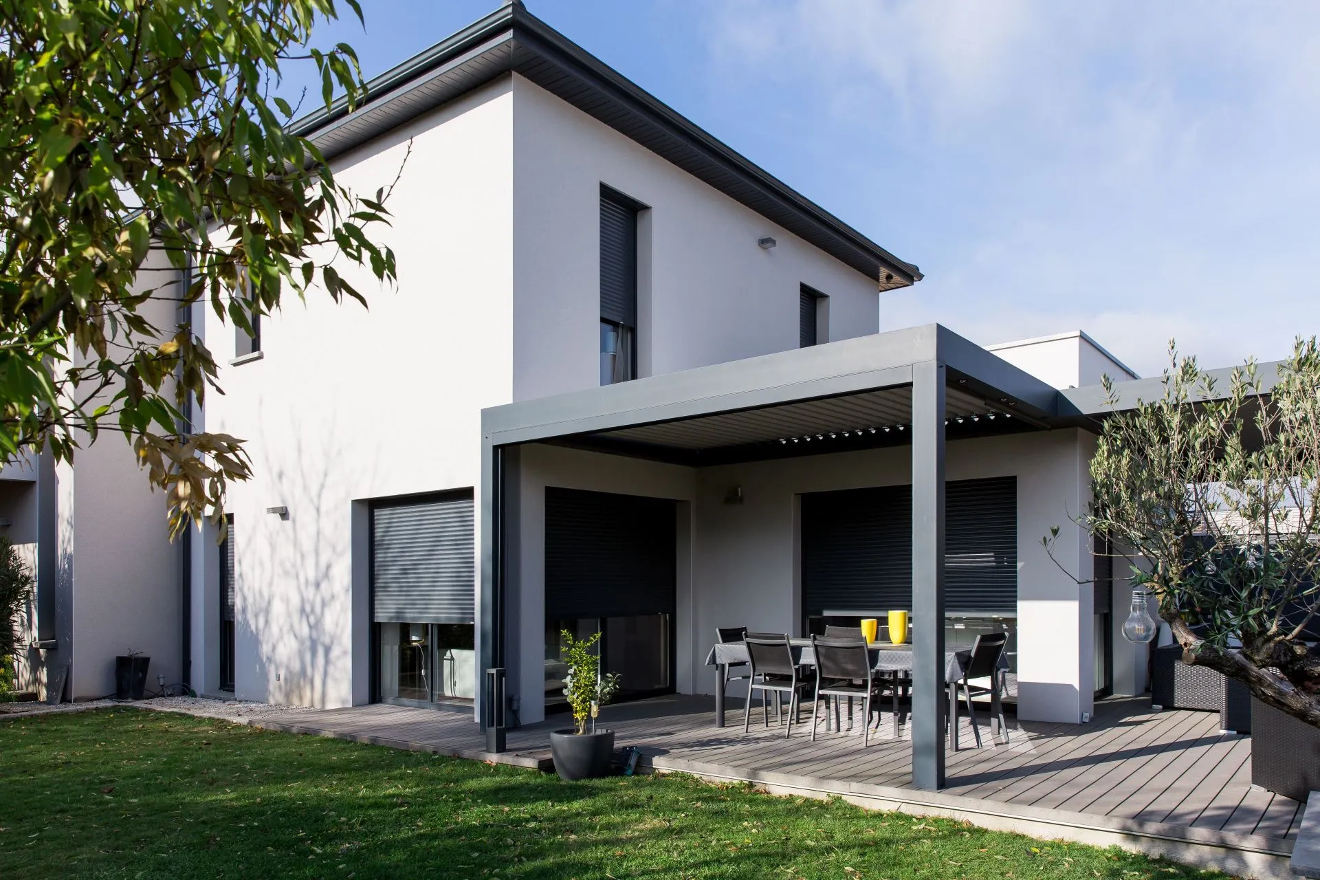 A modern two-story house with white walls and dark window shutters. The house features a covered patio area with a dining table and chairs. The patio has a pergola-style roof. The surrounding area includes a grassy lawn and a few potted plants. The sky is clear and sunny.