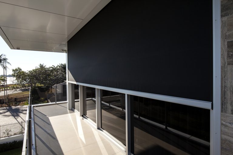 The photo shows a modern building with a large black exterior roller shade covering the windows on an upper floor. The shade is extended, providing shade to the balcony area. The building has a sleek design with a flat roof and glass windows. In the background, there are trees and a glimpse of a waterfront.
