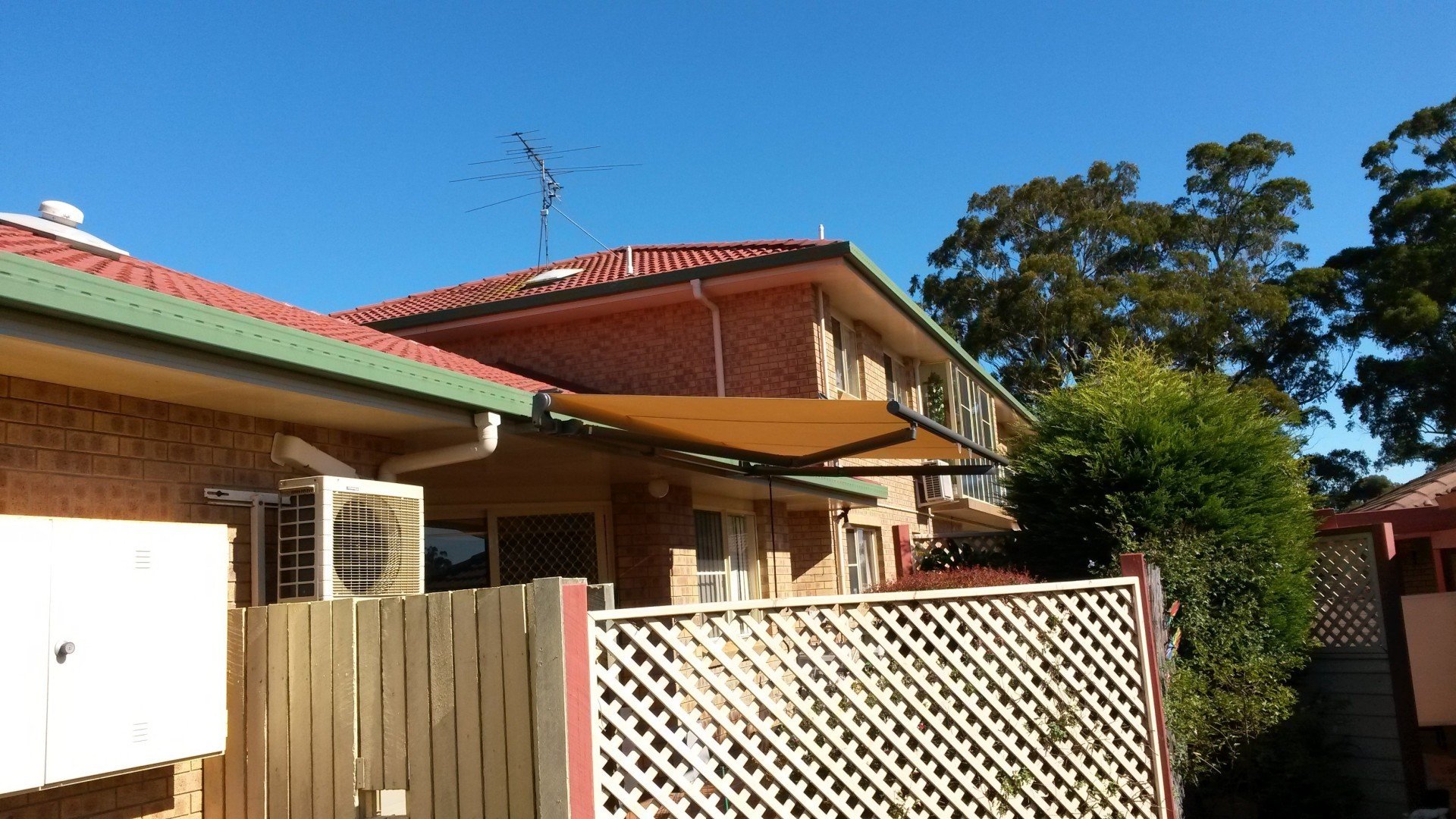 A two-story brick house with a red-tiled roof is shown. The house has a green gutter and a beige shade sail extending from the side. An air conditioning unit is mounted on the wall. A lattice fence is in the foreground, and there are trees in the background under a clear blue sky.