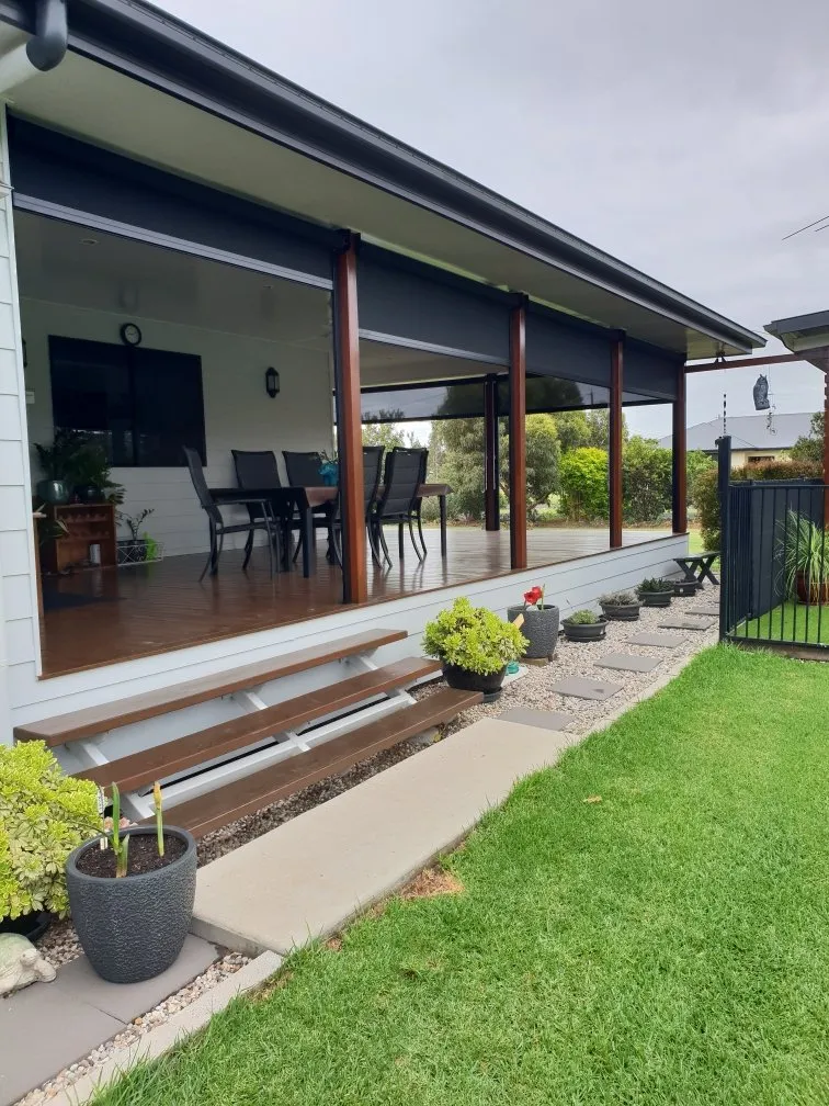 This image shows a modern outdoor patio area attached to a house. The patio is covered and features a dining table with chairs. There are potted plants along the edge of the patio and steps leading down to a well-maintained lawn. A fence is visible on the right side, and the sky appears overcast.
