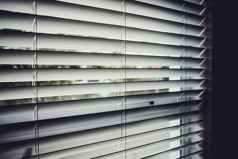 The image shows a close-up of horizontal window blinds, partially open, allowing some light to filter through.