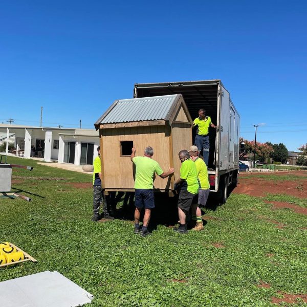 Four people are unloading a small wooden structure from a truck onto a grassy area. They are wearing bright yellow shirts, and the sky is clear and blue.