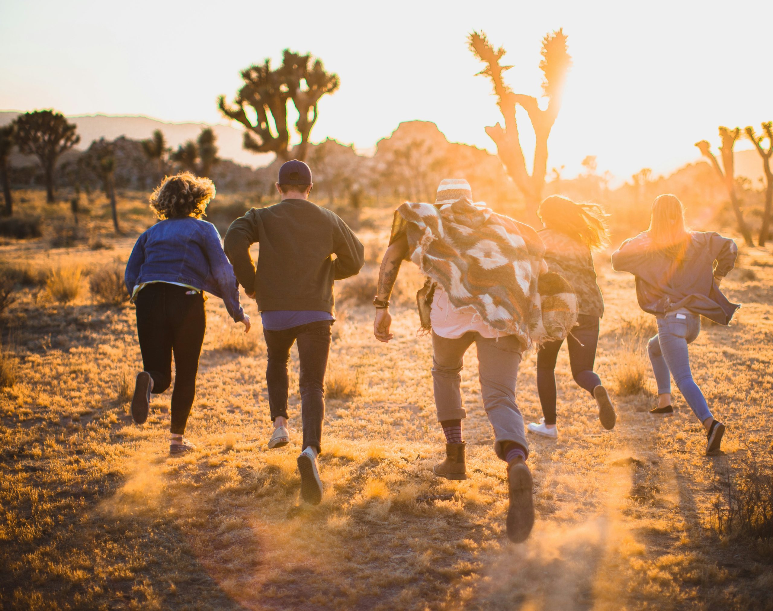 A group of people running through a desert landscape at sunset. The sun is low in the sky, casting a warm glow over the scene. The ground is sandy, and there are sparse desert plants and Joshua trees in the background. The people are wearing casual clothing and appear to be enjoying the moment.