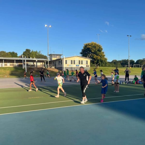 A group of people, including children and adults, are playing on an outdoor sports court. The court is surrounded by grass and trees, with a building in the background. The sky is clear and sunny.