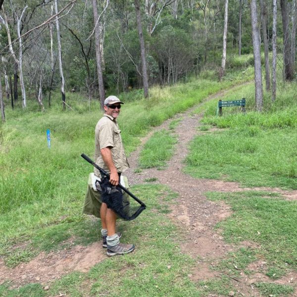 A person in outdoor attire stands on a dirt path in a grassy, wooded area. They are holding a large black object and looking back towards the camera. The path leads into a forested area with tall trees. The image has a "Koala" logo in the bottom left corner.