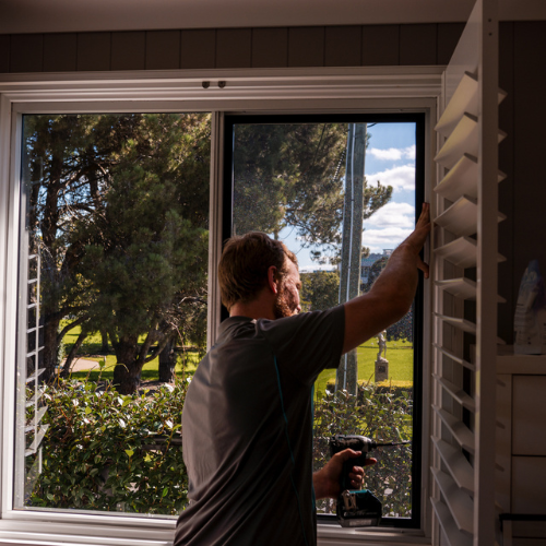 A person is installing or adjusting white plantation shutters on a window. They are using a power drill, and the window is open, showing a view of trees and a grassy area outside.